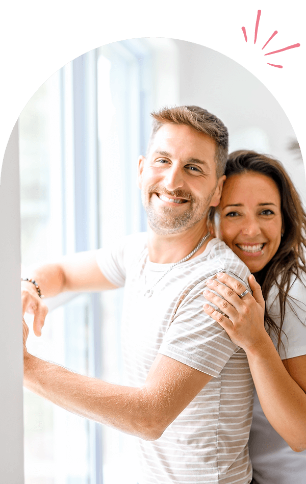 Group 1321317415 (1) A happy couple smiling and embracing indoors near a window.
